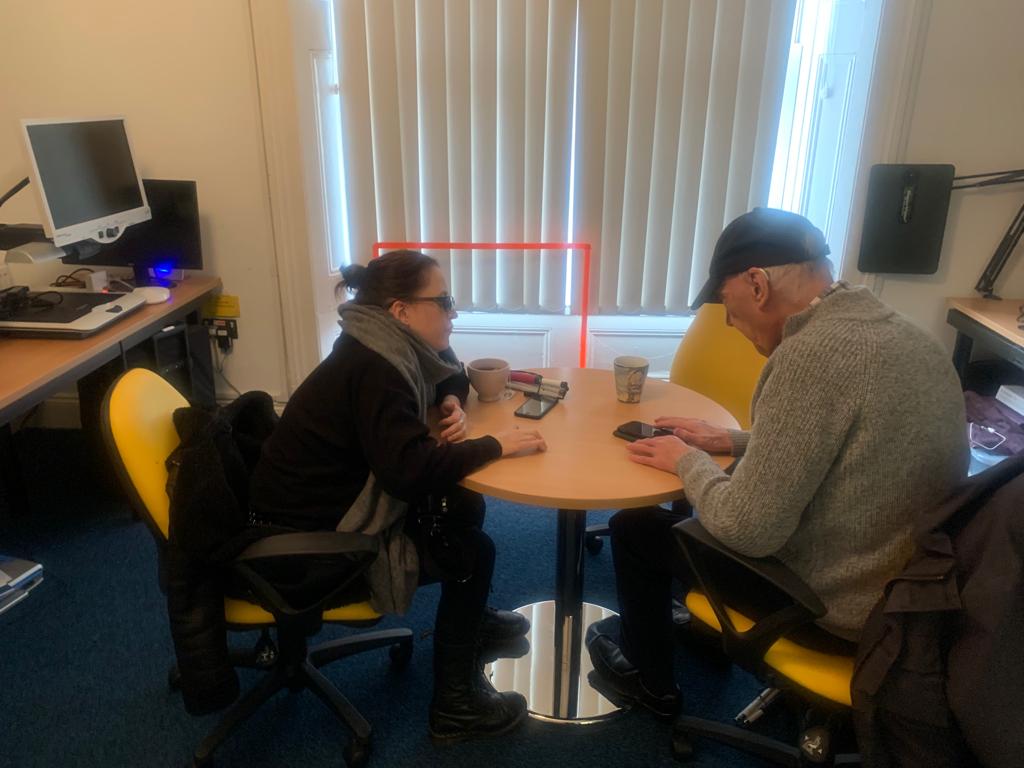 Richard sitting at a desk in the Karten suite, learning how to use his iPhone from volunteer Hana who is also visually impaired. 