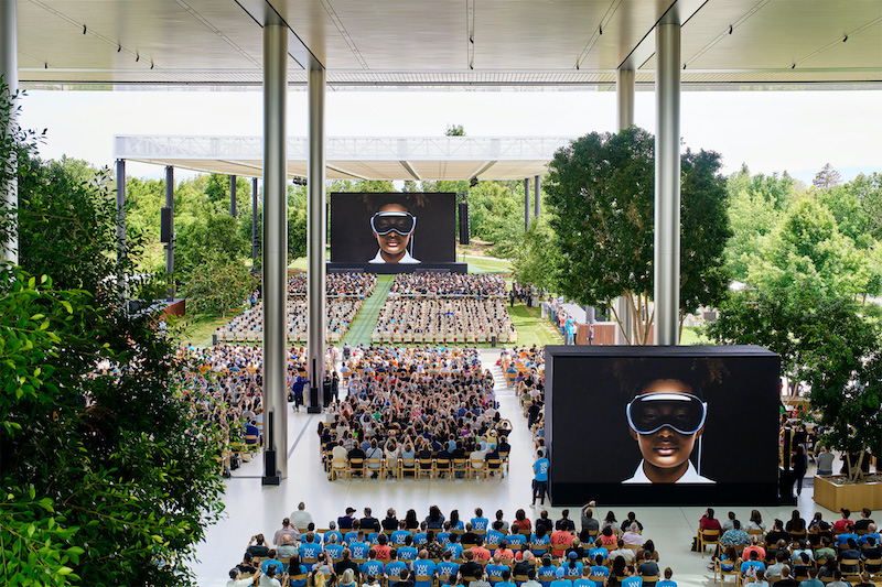 Hundreds of developers sit in chairs at Apple Park watching the WWDC23 keynote.Two large screen display an image of the Vision Pro