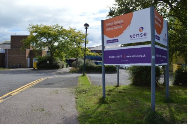 The entrance to Sense College  in Loughborough, with a large sign displaying the name, logo, contact details. The background includes a curved road, brick buildings, trees, and parked cars under a partly cloudy sky.