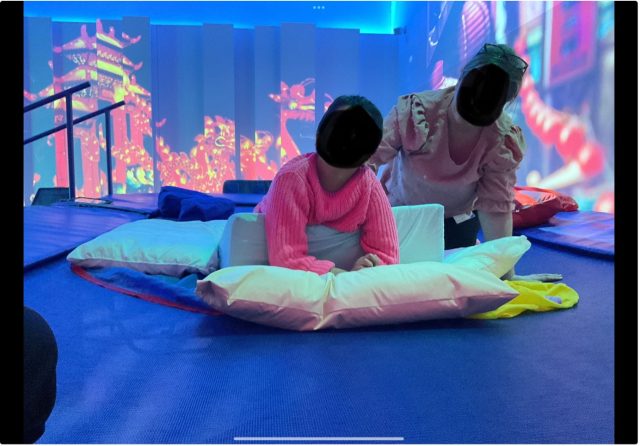 oung person lying on a therapy wedge during a rebound session, supported by a staff member in a sensory 4D suite, with colourful cityscape projections in the background.