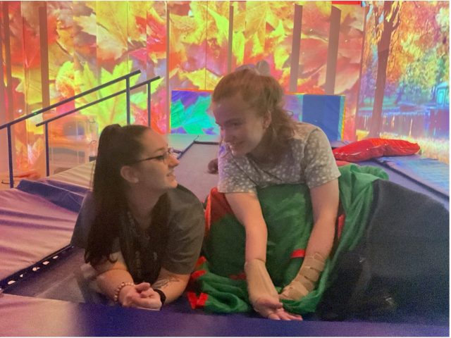 Two young women smile at each other while lying on a padded surface in a sensory 4D suite, with autumnal forest projections creating an immersive background.