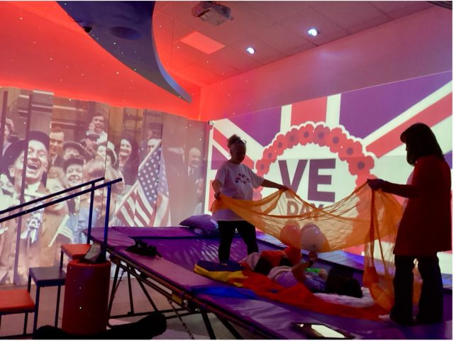 Two staff members support a young person on a trampoline during a VE Day-themed rebound therapy session in the immersive 4D suite, with vibrant projected visuals of the Union Jack and historical photos in the background.