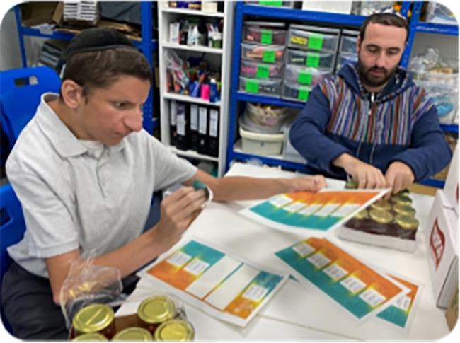 Two members of the Enterprise Team sorting and packaging colourful cards at a workstation, surrounded by storage shelves filled with supplies.