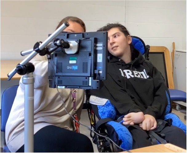 A student using a communication device with eye gaze technology, supported by a staff member, preparing for their turn in the Sense College Rap Battle.