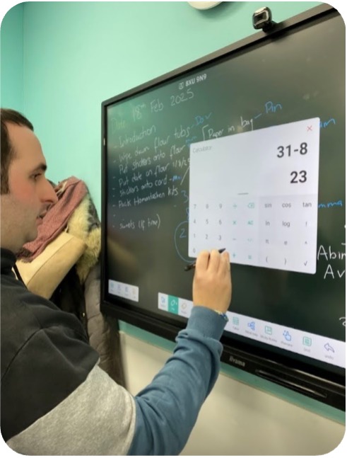 A learner interacts with a touchscreen display, using an on-screen calculator app while supervised by a support staff member.