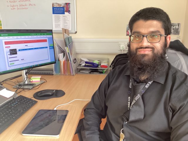 Waj, a former student, smiling at his desk in his new role as a Level 3 apprentice with Everyone Learning, seated beside a computer and tablet.