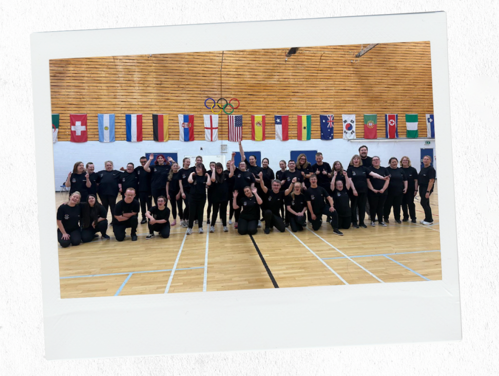 Large group of Buzz Hub performers posing and smiling in a sports hall beneath a row of international flags and Olympic rings.