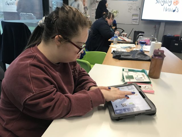 Trish using an AAC app on a tablet to communicate while seated at a table in a group learning session.