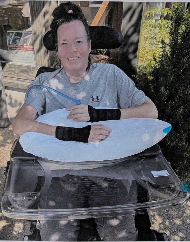 Julia, smiling seated in a powered wheelchair outdoors, with supportive arm rests and a lap tray.