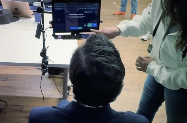 A person is seen from behind seated at a table, facing a tablet mounted on a stand. Another person stands beside them pointing at the tablet screen, as if demonstrating an assistive technology setup in a workshop setting.