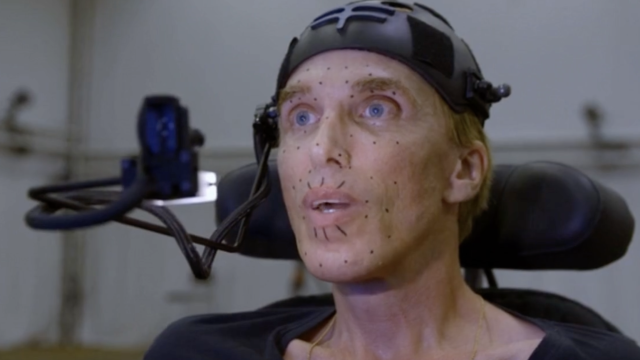 Close-up of a person using assistive technology in a lab setting, wearing a sensor cap and facial tracking markers, seated with head support, with a microphone or camera arm positioned beside them.