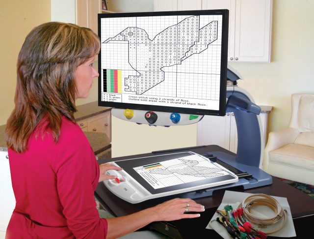Person using a desktop electronic magnifier (CCTV) to view an enlarged cross-stitch pattern displayed on a large screen above the reading surface.