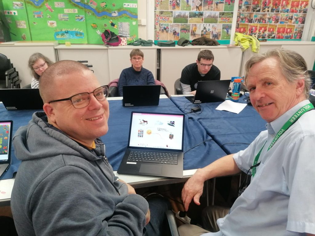 Five people sit around a large table covered with a blue cloth in a bright classroom. In the foreground, two men smile toward the camera — one wearing a grey hoodie, the other in a light blue shirt with a green volunteer lanyard. Behind them, three other group members work quietly on laptops. Colourful displays and photographs cover the walls in the background.