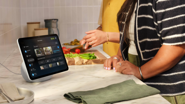 A woman prepares food at a kitchen worktop while an Amazon Echo Show smart display in the foreground shows the Alexa+ Smart Home dashboard, displaying camera feeds, home updates, and device controls.