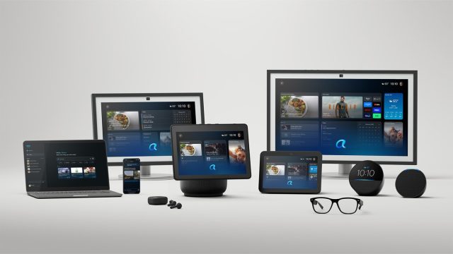 A product family photograph showing the Amazon Alexa+ ecosystem across multiple devices, including Echo smart speakers, Echo Show smart displays, a large screen display, a laptop, a smartphone, smart glasses, and earbuds, all arranged on a white surface and displaying the updated Alexa+ interface.