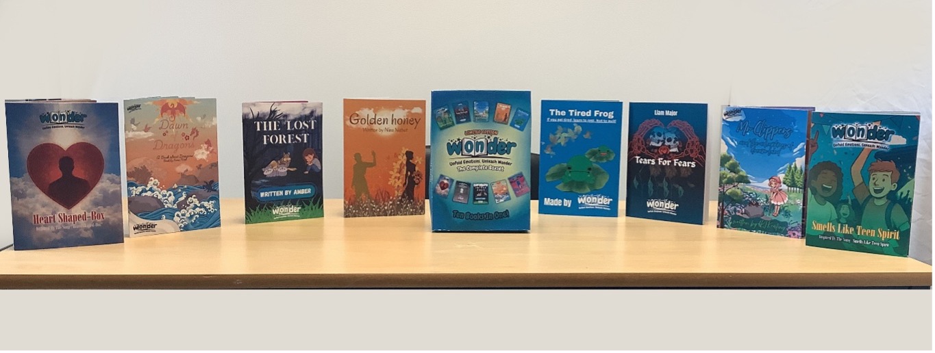 A row of nine student-authored books published by Wonder, displayed on a wooden table. Titles visible include Heart Shaped-Box, Dawn Dragons, The Lost Forest, Golden Honey, The Tired Frog, Tears For Fears, and Smells Like Teen Spirit. In the centre is a limited-edition Wonder box set titled "The Complete Boxset — Ten Books in One."