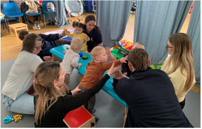 A group of adults and young children sit together on the floor in a bright, welcoming room. A therapist or practitioner engages closely with the children while parents observe and participate. Colourful toys and soft furnishings are visible in the background.