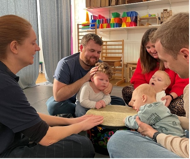 Four adults sit closely together on the floor, each holding or interacting with a baby. The group faces inward, creating an intimate circle. The setting appears warm and relaxed, with colourful resources visible in the background.