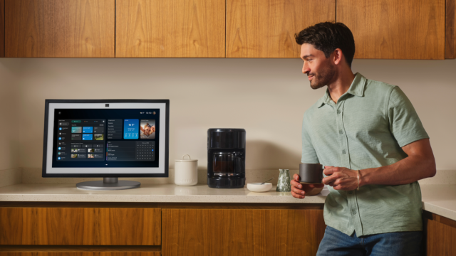 A man holding a mug stands in a modern kitchen, smiling as he looks at an Amazon Echo Show large screen display on the worktop beside a coffee maker. The screen shows the Alexa+ home dashboard interface.