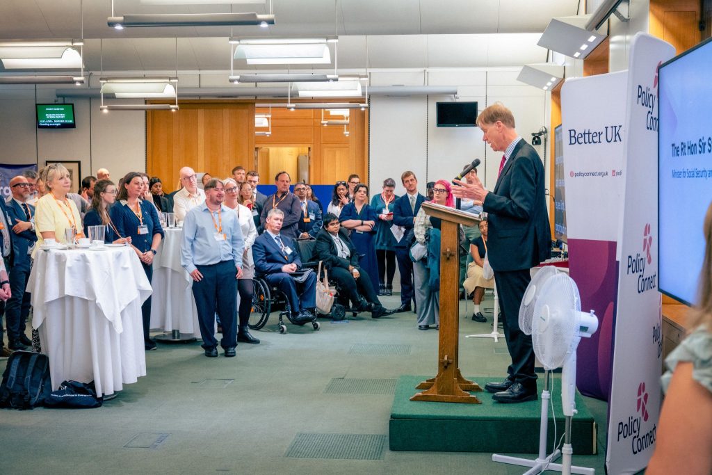 Rt Hon Sir Stephen Timms MP speaks from a lectern at Portcullis House, addressing an audience that includes several wheelchair users, with a Policy Connect screen displaying his name in the background.