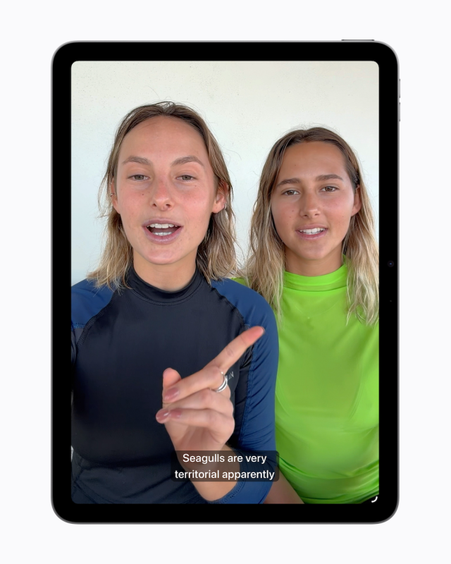 iPad showing a personal video of two women in swimwear, one gesturing while speaking. A generated caption reads 'Seagulls are very territorial apparently' automatically overlaid at the bottom of the video.