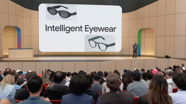 A presenter stands on stage at Google I/O 2026 in front of a large screen reading 'Intelligent Eyewear,' showing two designs of smart glasses, one a sunglasses style and one a clear-lens style, with an audience seated in the foreground.
